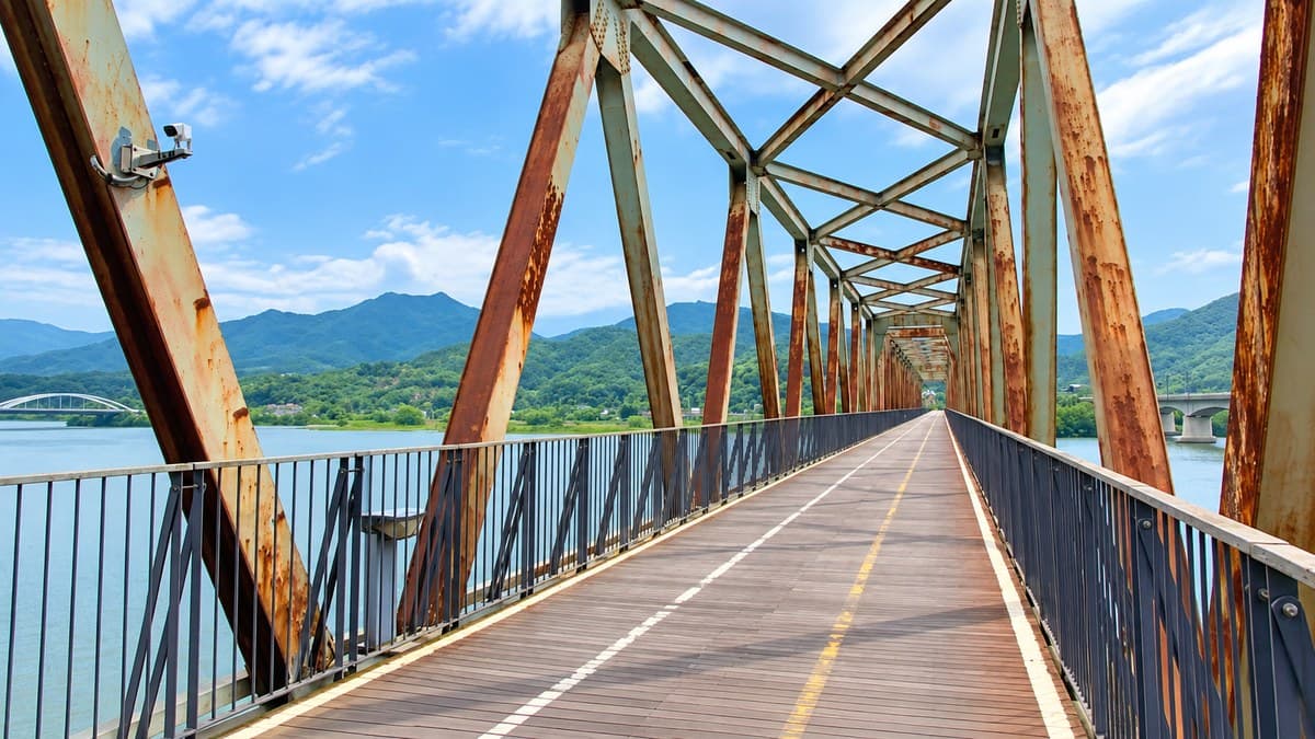 A cycle path running along the deck of a converted steel railway bridge over a wide river, with rusted trusses overhead and green mountains beyond