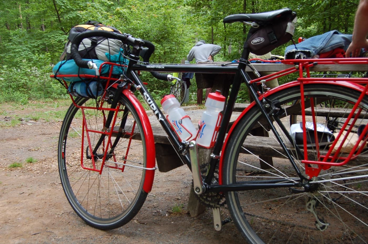 A loaded touring bicycle with front and rear racks, panniers, and water bottles, parked at a trailside picnic table