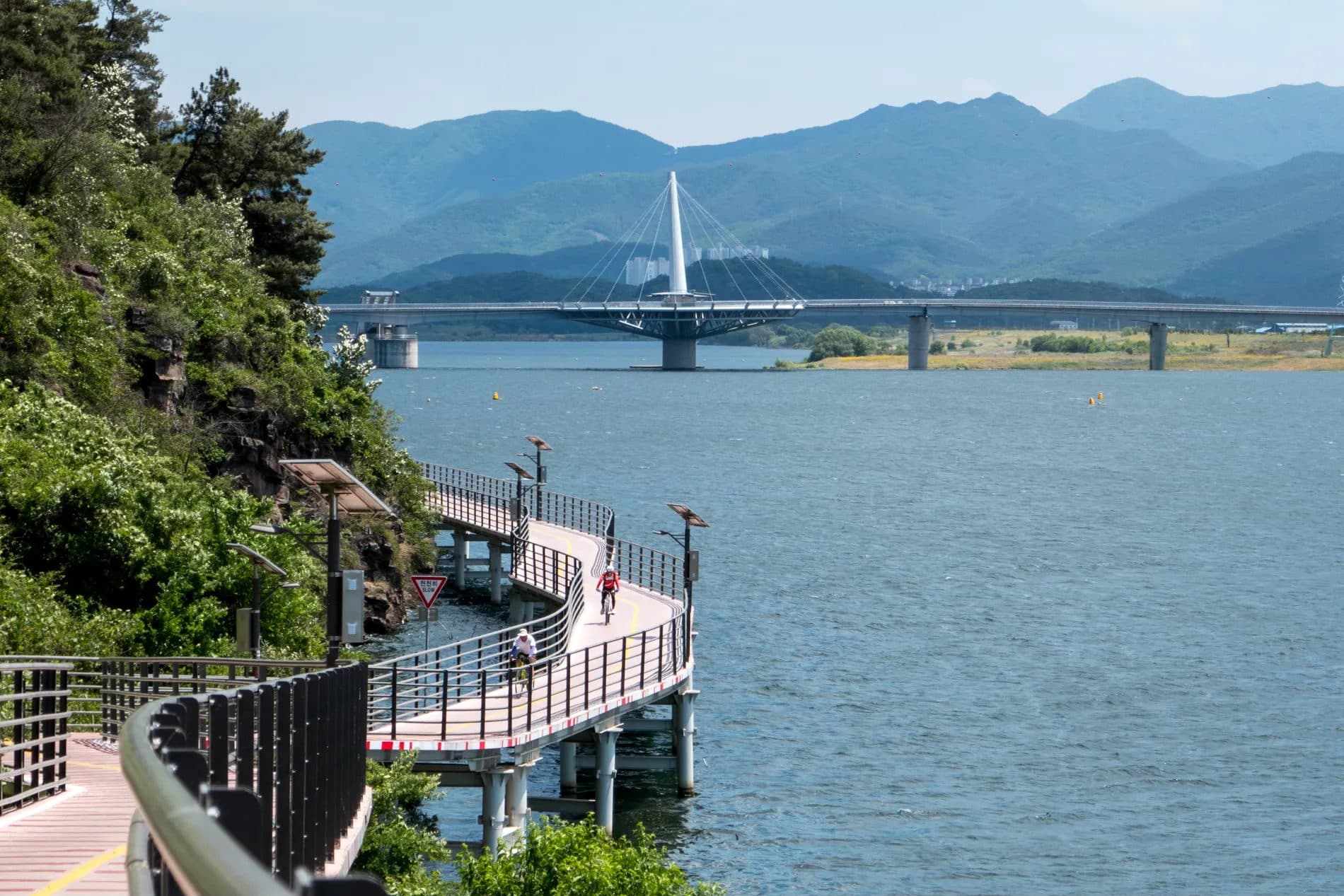 Two cyclists on a boardwalk section of the path built out over a wide river, curving toward a white cable-stayed bridge with mountain ridges behind it