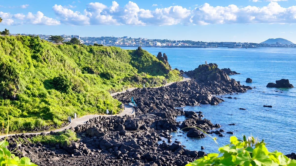A narrow coastal path threading through black volcanic rock on Jeju, green headland on one side, blue sea on the other and Jeju City along the shore behind